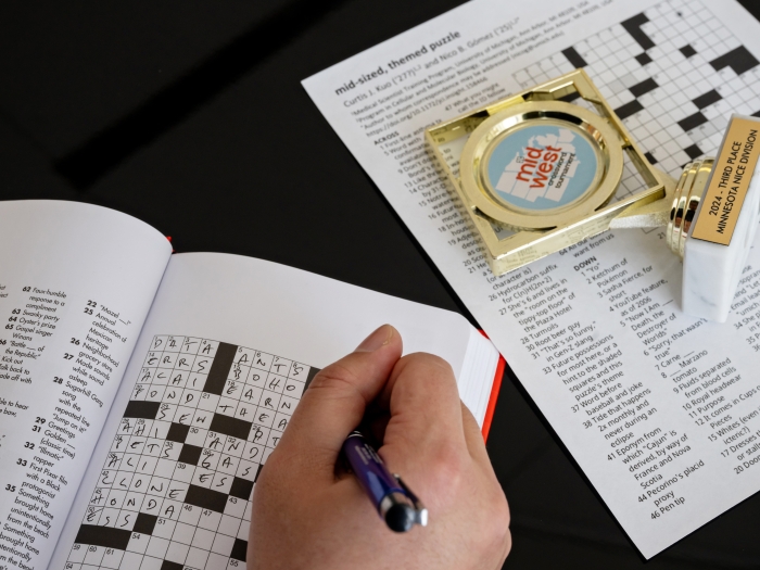 A person filling out a crossword puzzle. Only their hand is visible. There's also a small trophy on the right side that commemorates winning a prize at a crossword puzzle competition.