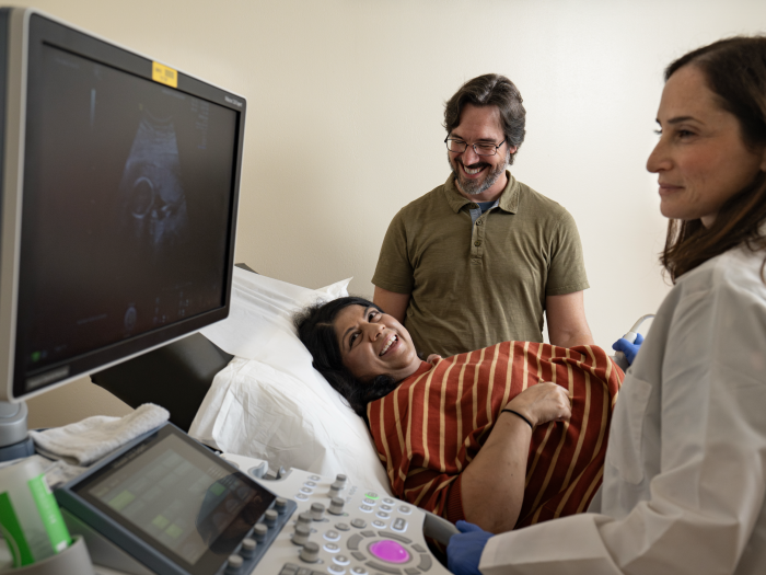 A smiling pregnant woman and her partner look at an ultrasound image that a doctor is showing them.