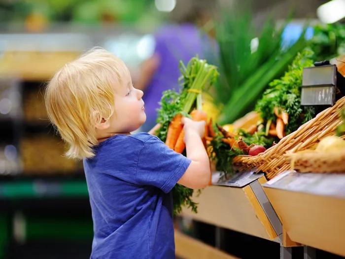 A small child in a blue t-shirt picks up a bunch of carrots from a produce display.
