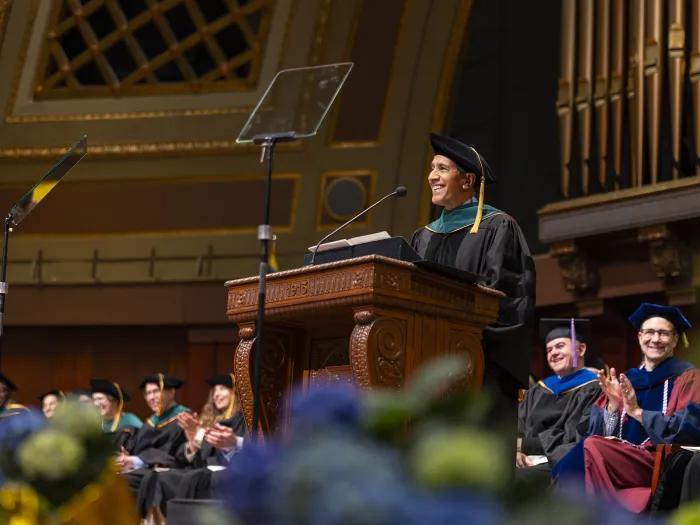 A speaker in academic regalia stands at an ornate wooden podium during a commencement ceremony, with seated faculty applauding nearby and organ pipes in the background.