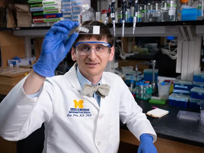 Lev Prasov, M.D., Ph.D., wearing a white coat, blue gloves, and safety glasses, views a microscope slide he is holding toward the viewer.
