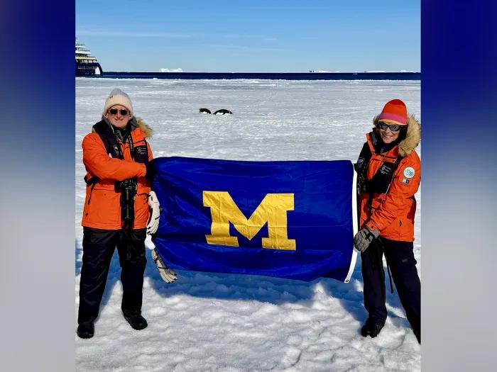 Bob and Debbie Merion, wearing orange parkas, stand on the Antarctic ice holding a U-M flag between them.