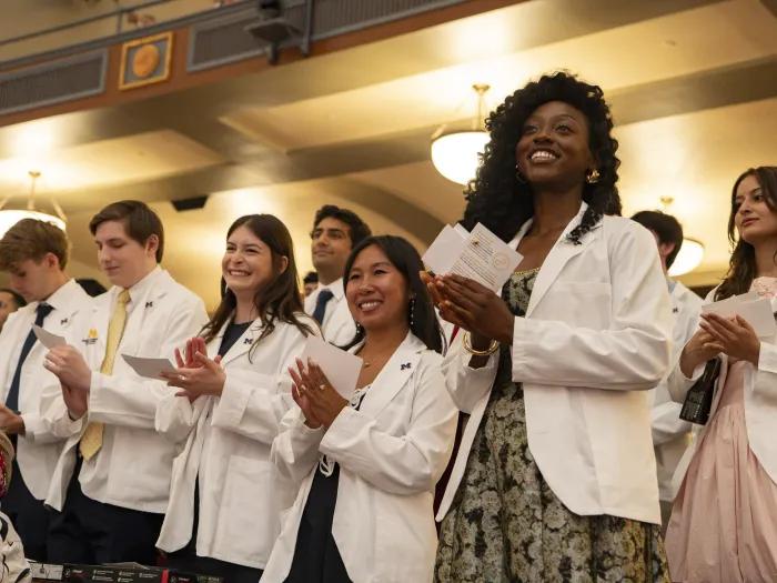University of Michigan medical students in white coats stand and applaud during a ceremony in an auditorium.
