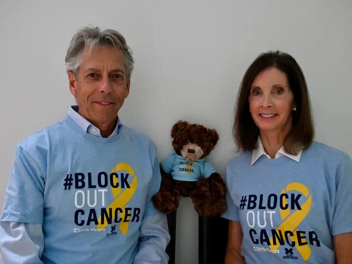 Andy and Linda Jacob smile at the camera beside a teddy bear, all wearing light-blue “#Block Out Cancer” shirts with yellow awareness ribbons.