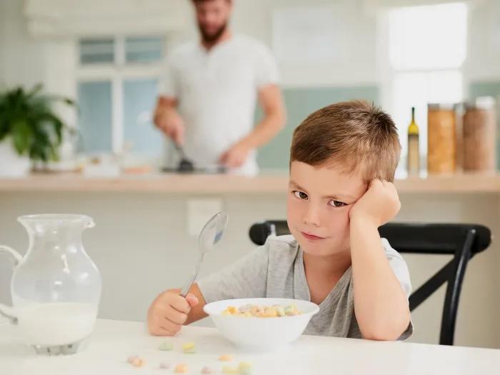 kid at table not wanting to eat breakfast with parent blurred in background white coloring milk on table