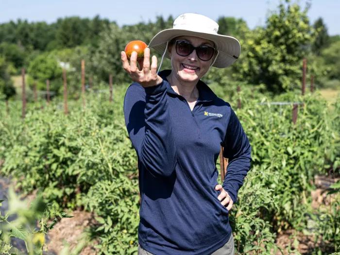 woman in michigan sweatshirt zipped up holding tomato smiling with hat on in green farm