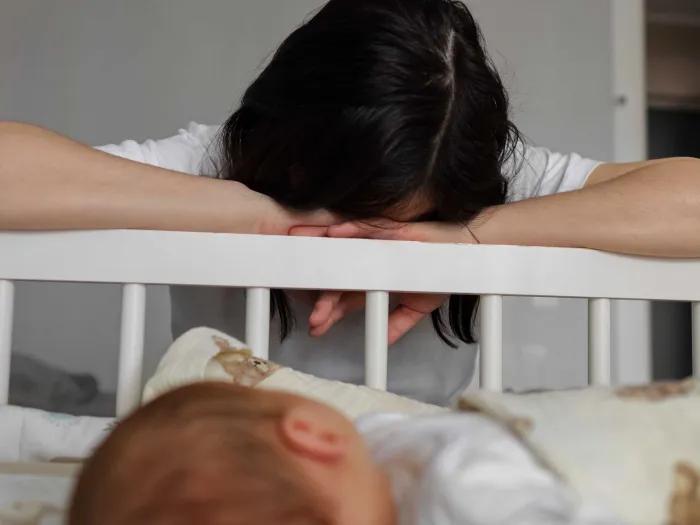 A dark haired woman with her head resting on her arms face down on the rail of a crib with a baby visible in the foreground