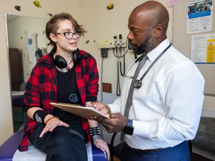 A photo of an African American male doctor showing a clipboard to a young gender non-conforming, trans, gay, pagan man.