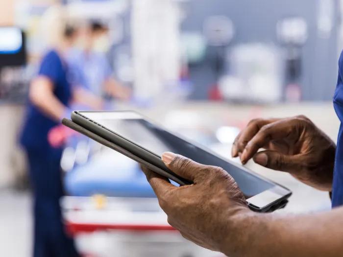 Two people in medical scrubs are in a blurred hospital setting. In the foreground, hands are holding a digital tablet.