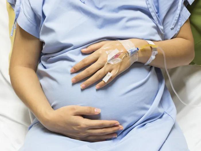 Woman's torso in a blue hospital gown, with her hands holding a pregnant belly and an IV in place.