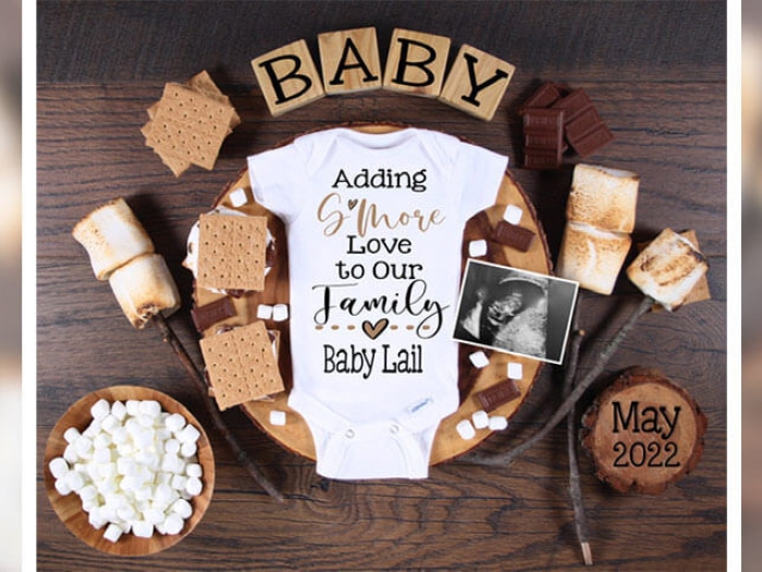 Baby onesie surrounded by smores, marshmellows and chocolate, reading adding smore love to our family baby Lail 