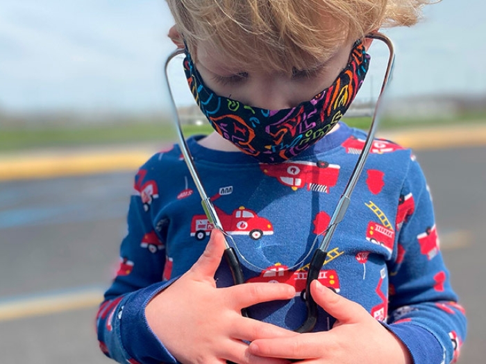 child looking down at stethoscope on his chest while wearing mask outside and wearing blue shirt with firetrucks on it