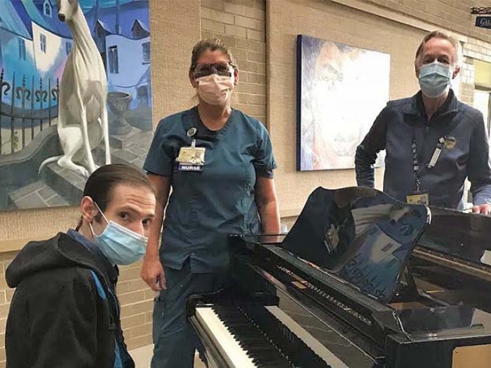 man sitting at piano with mask on and nurse with mask on standing near him and man with mask on standing across piano