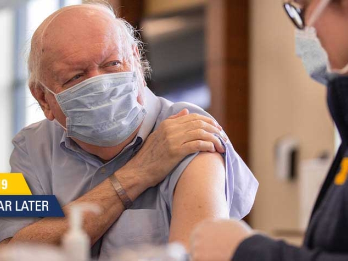 older man with white hair and blue face mask on lifting sleeve on blue shirt to get vaccine from worker