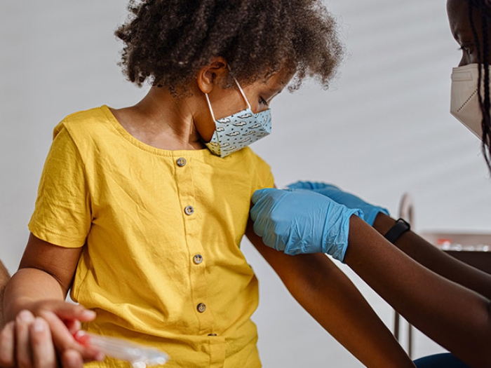 child in yellow shirt sitting looking down on arm with blue mask on and nurse with globes putting band-aid on
