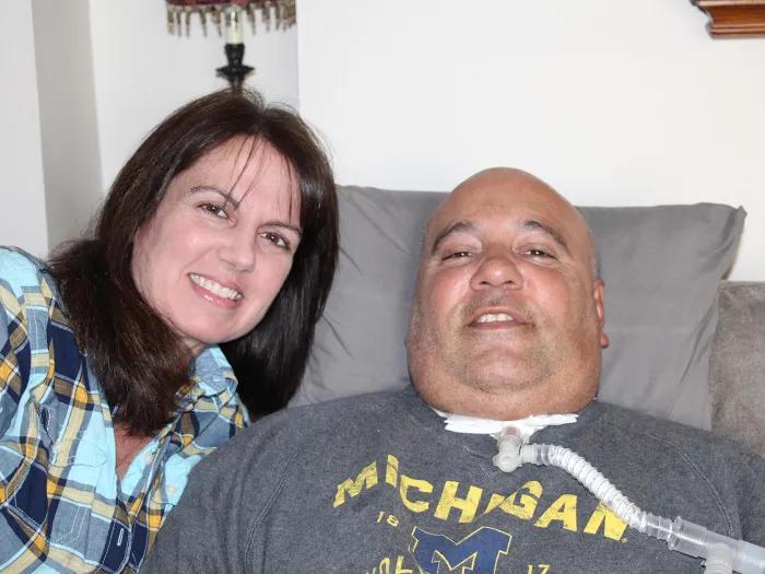 woman with dark hair standing next to man with tracheostomy sitting in chair