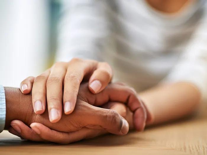 Two hands rest gently on one another on a wooden table, conveying support and compassion.
