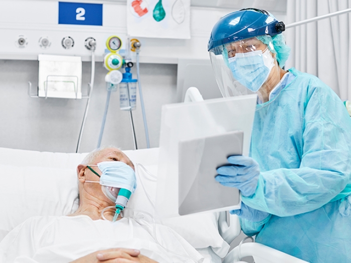 older patient in white hospital gown in ICU with doctor in blue protective gown and equipment and face shield holding ipad up