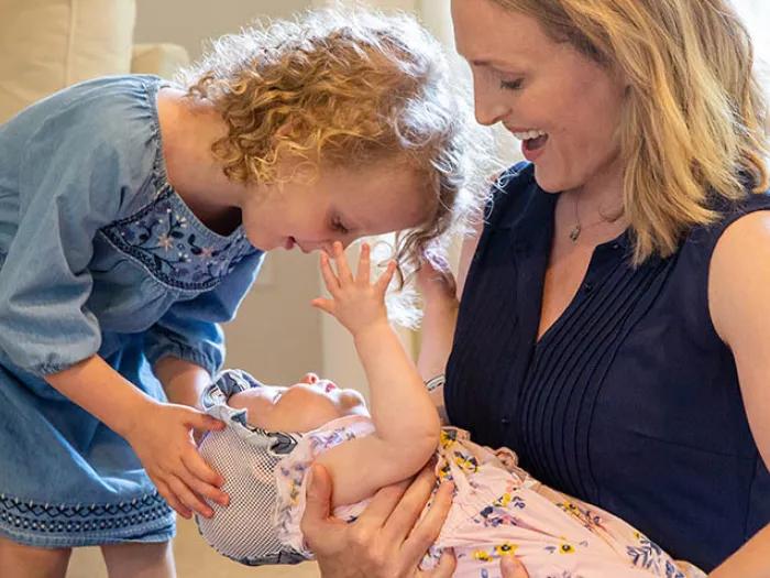 Mother in blue sleeveless top holding and look down at her baby with blue hat and pink outfit with flowers and toddler also looking at baby wearing blue dress