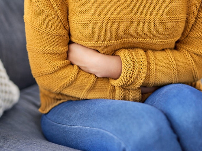 woman holding stomach pain mustard colored shirt jeans sitting on couch