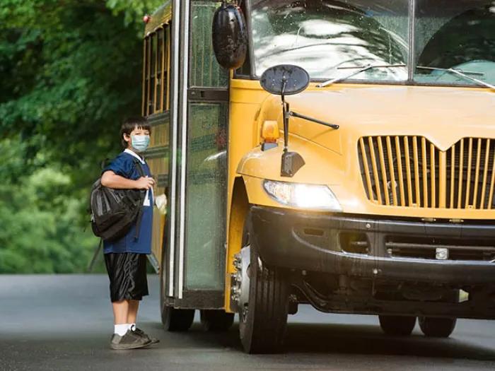 Child with brown hair and mask on in front of open doors of school bus wearing backpack