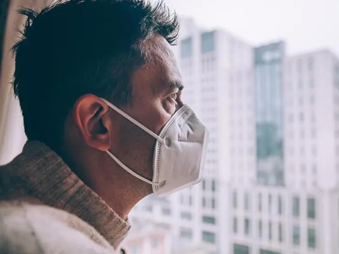 Man with mask on staring outside the window looking at city.