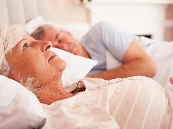 Elderly woman lying awake in bed next to a sleeping man, looking contemplative.