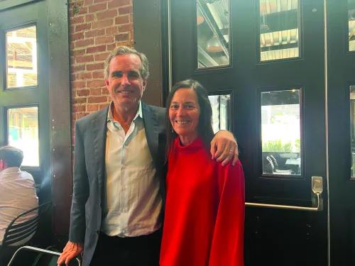 A man and a woman stand next to each other smiling at the camera. The man, wearing a suit, has his arm around the shoulder of the woman, wearing a red sweater.