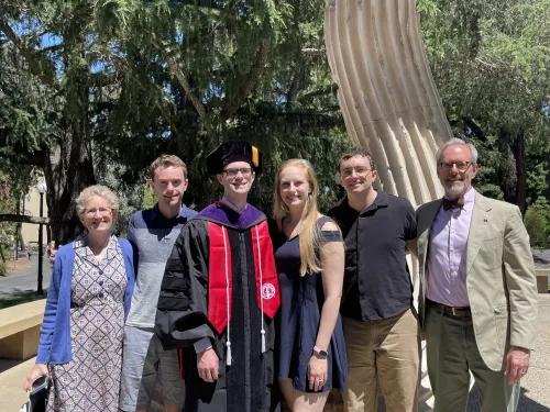 Group photo of six people. One of them near the middle is wearing a graduation cap and gown.