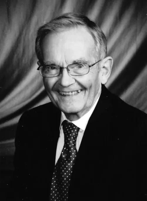Headshot of Lawrence Sweet. He is wearing a suit and tie and glasses. He is smiling and looking at the camera. In the background is a draped curtain.