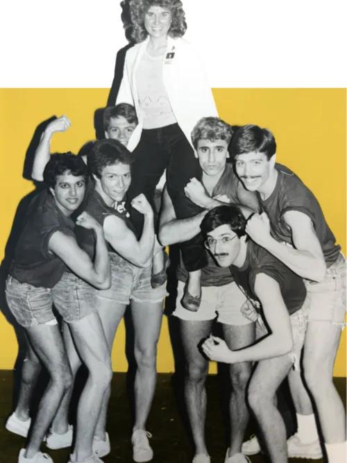 Six male medical students strike muscle poses while two hoist a female medical student above their shoulders. The men are in short shorts and T-shirts. The woman is wearing a white doctor's coat and slacks.