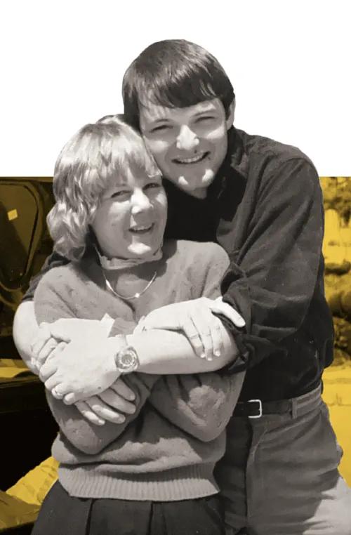 Black and white photo of two medical students embracing. They are both looking at the camera and smiling. They are set against a maize-washed background