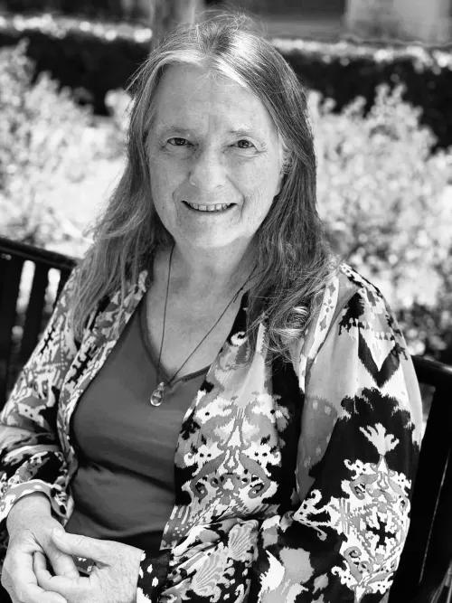 Black and white photo of Sue Boiko seated outdoors on a bench in a garden. She's wearing a print cardigan. She's smiling and looking at the camera.