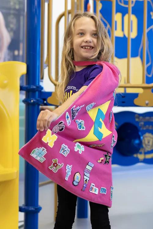 Photo of Evalynn standing in front of a play structure at a playground. She is wearing a pink cape with many patches on it and a large E over a lightning bolt.