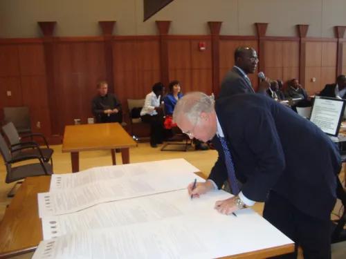 Side view of Johnson leaning over a table covered with four very large official-looking documents. He is signing one of them. Several people are standing or seated in the background and the setting appears to be a meeting or conference.