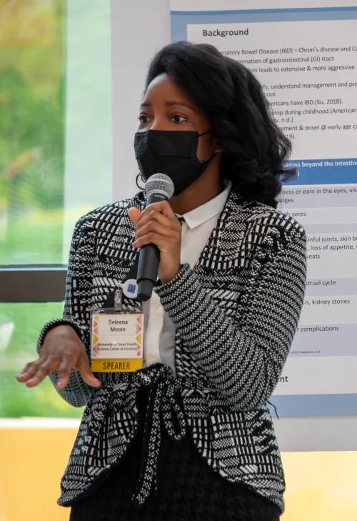 A speaker in a blazer and a mask speaks into a microphone in front of an academic poster.