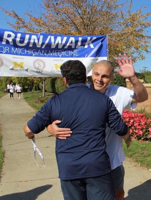 Giavino hugs his dad, Bassam, who smiles and waves toward the viewer. A large run/walk banner is in the background.