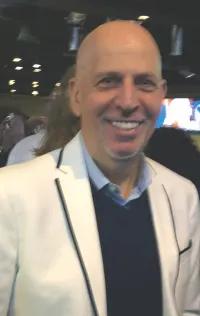 Man with bald head smiling at the camera. He's wearing a white jacket with a black sweater and blue button up shirt. 