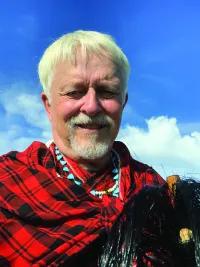 Man with white hair smiling at camera. He's wearing a red and black plaid shirt or shawl.