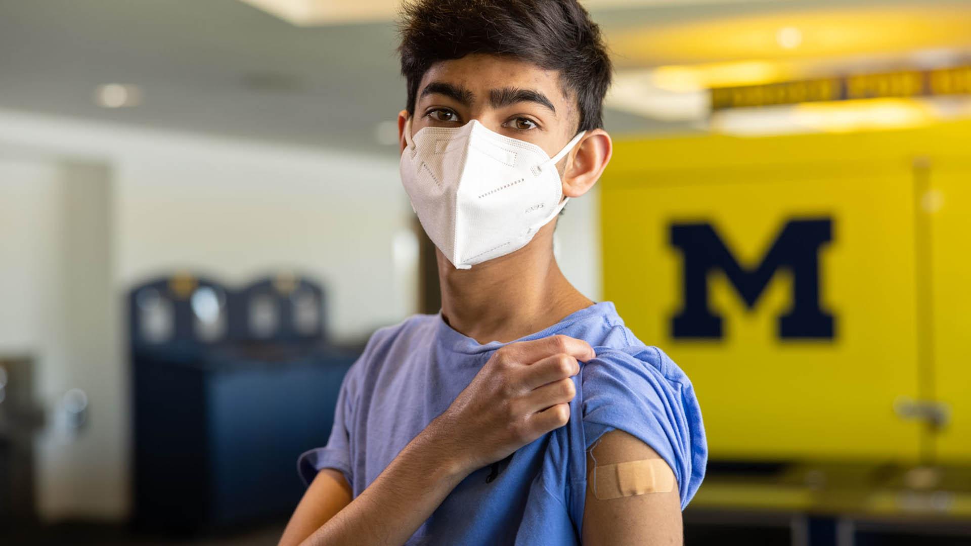 Teenage boy wearing mask and pulling up blue t-shirt to show bandage on upper arm