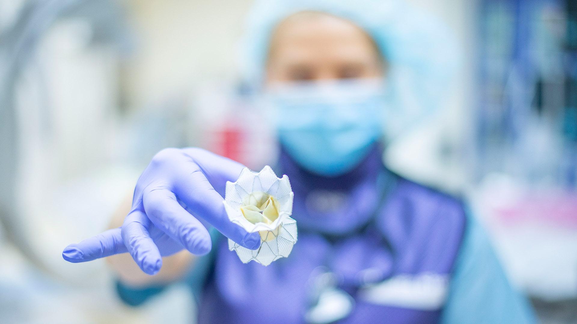 Flower-like TAVR device at forefront held by woman in pale blue surgical garb with lavender surgical glove and dark blue overgarment 
