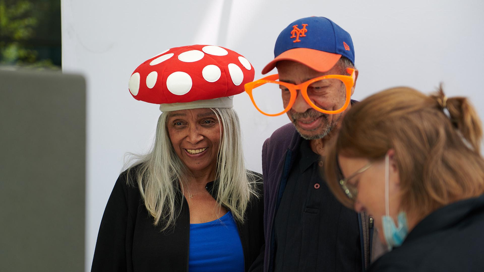Woman in red and white mushroom hat, and a man in orange oversized glasses smile.