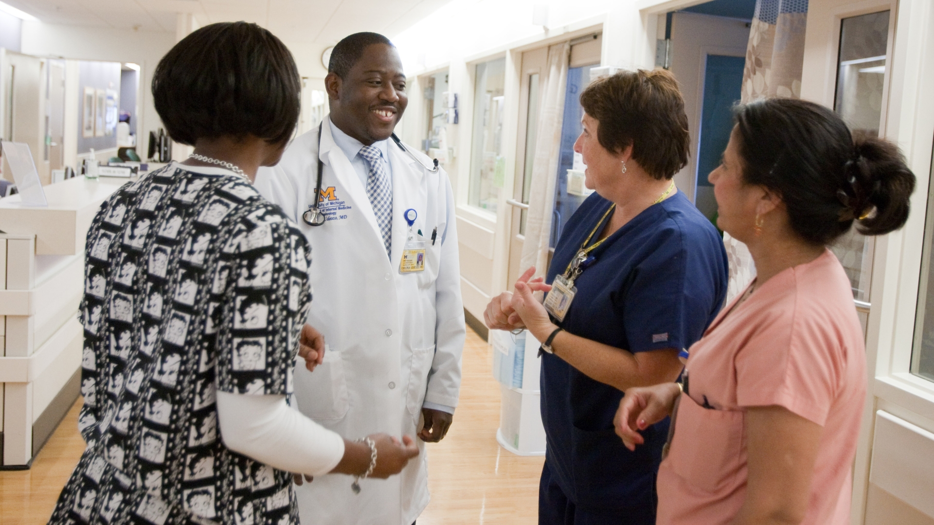 Black male doctor wearing white coat talking in hospital hallway to three diverse female medical professionals