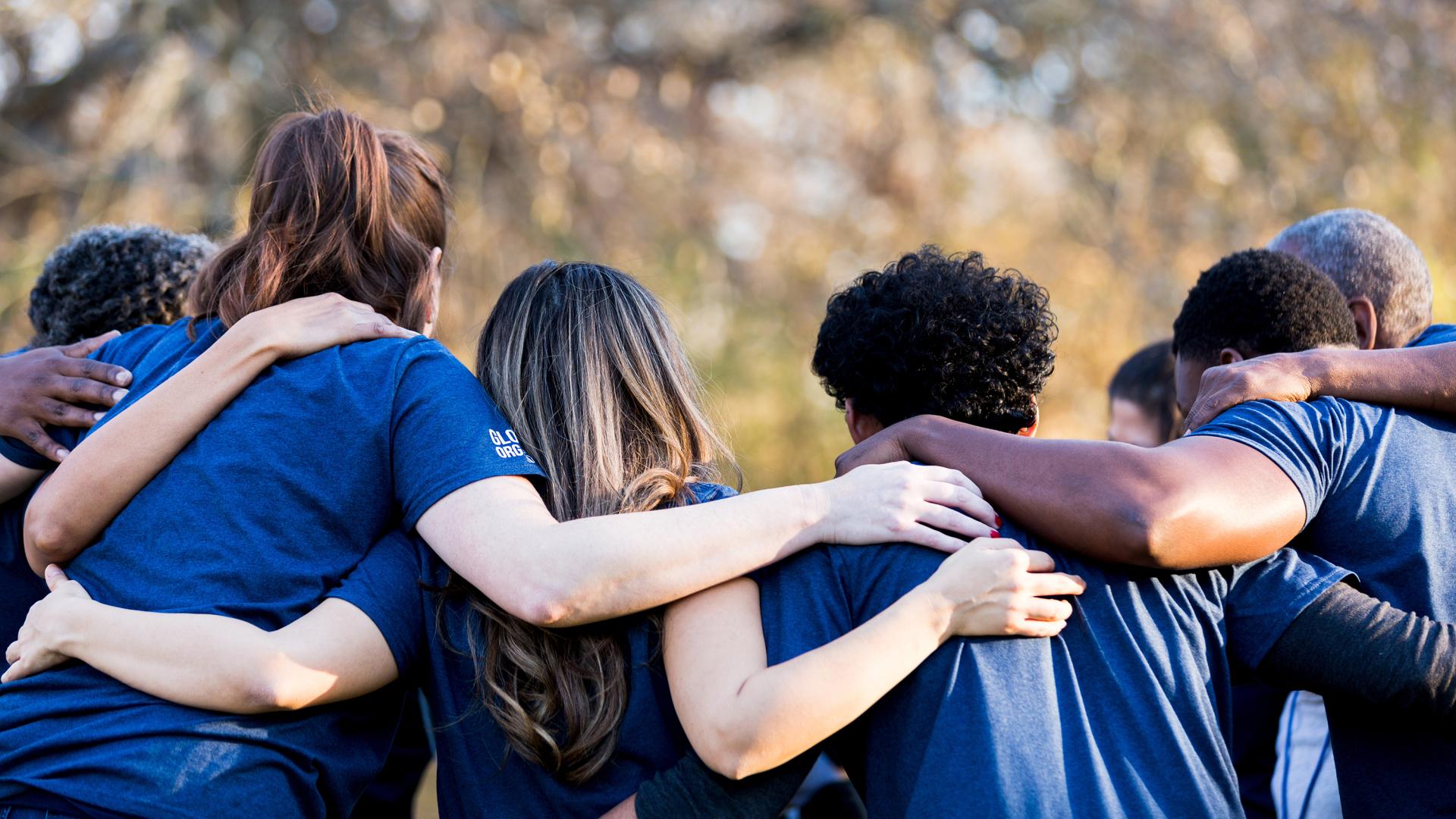 Diverse group of people wearing blue shirts shown from behind with arms around each other