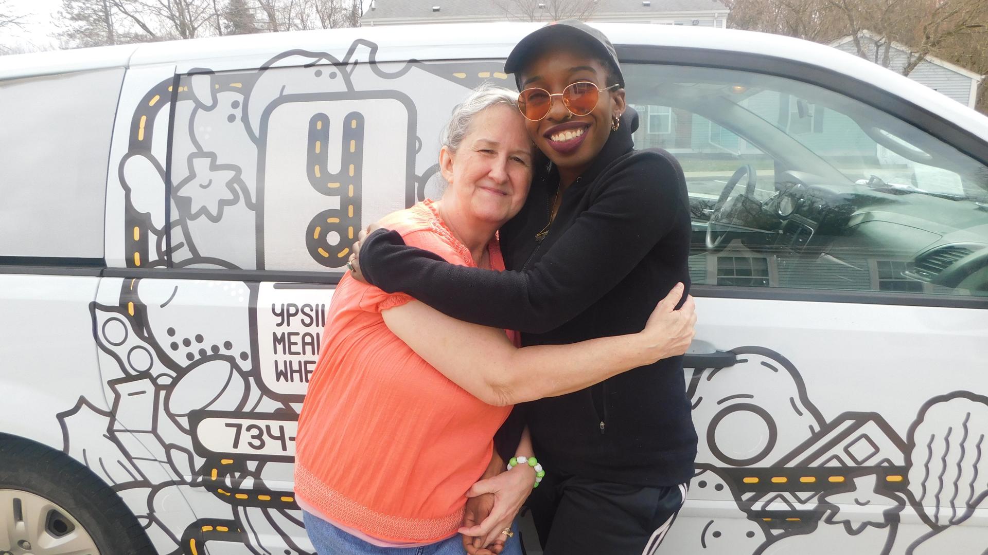 Two women hugging with Ypsi Meals on Wheels truck in background