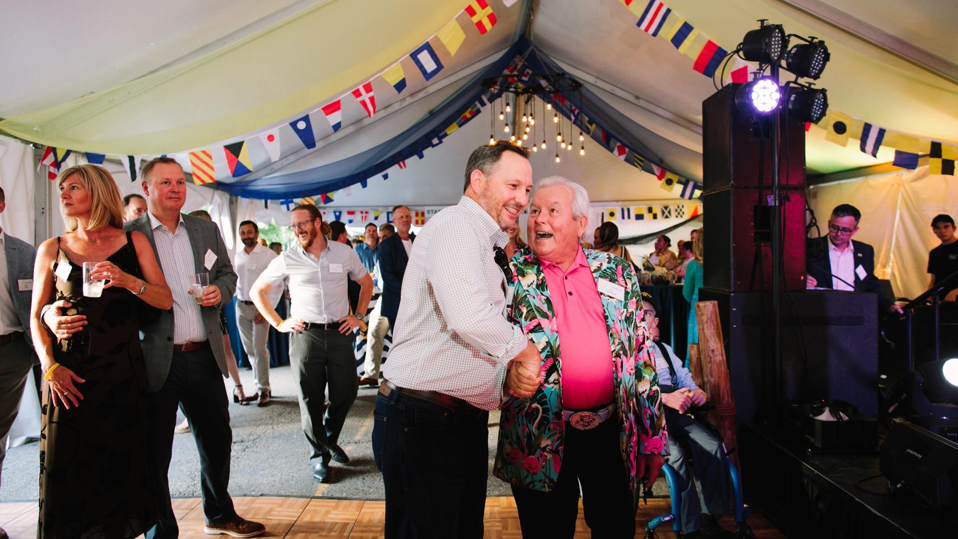 Man with pink shirt and colorful jacket shaking hands with another Event on Main attendee under a tent with nautical decorations