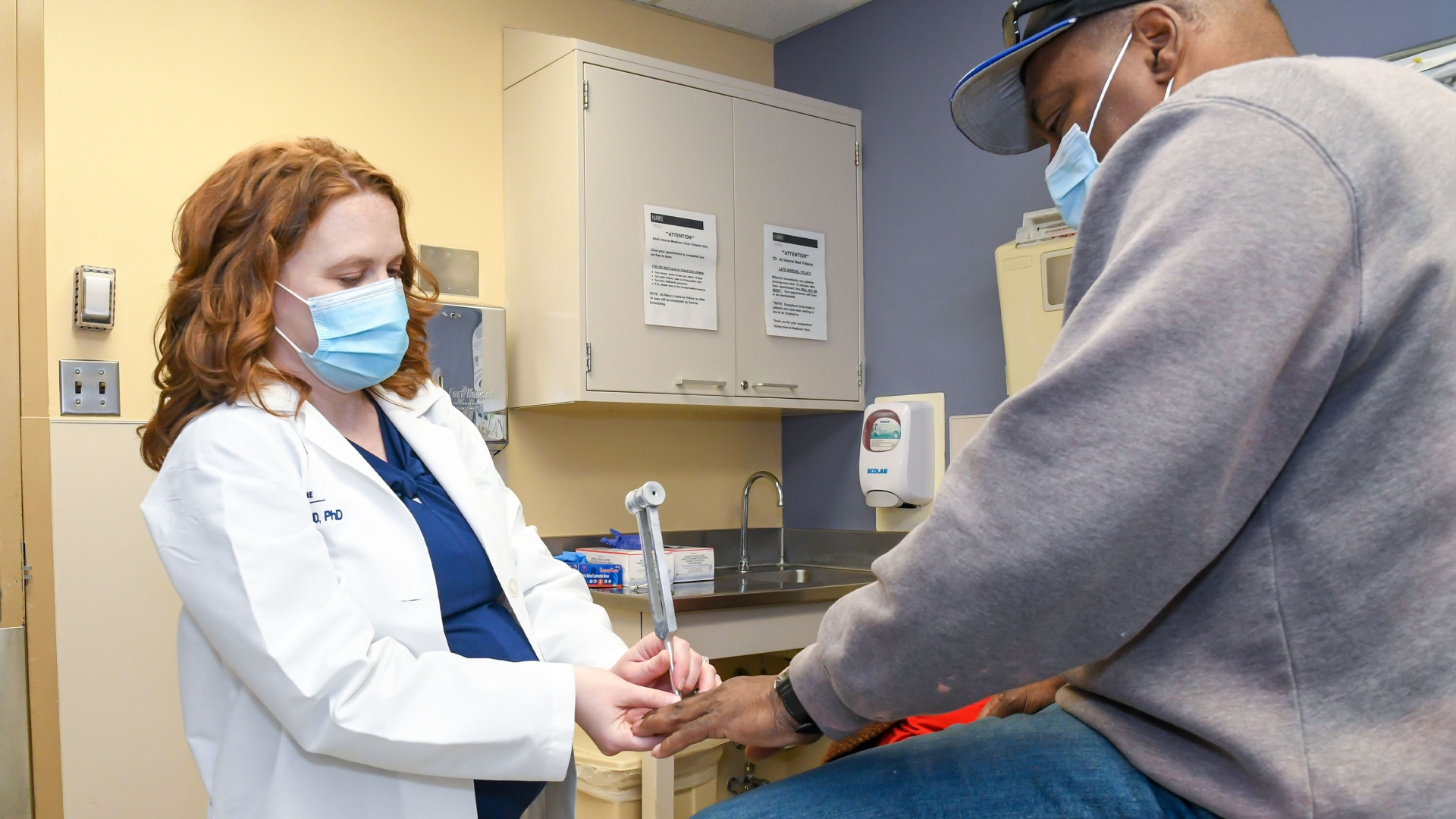 doctor in white coat with dark blue scrubs touching hand of patient in grey sweater and baseball cap in exam room 