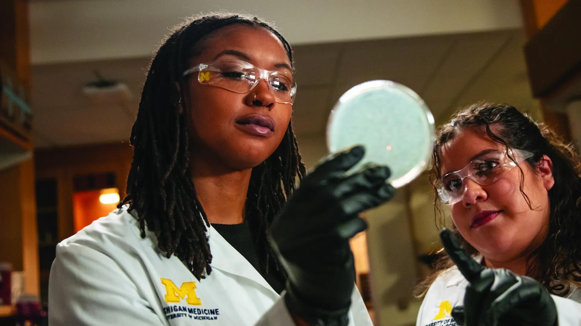 Two researchers in white coats and safety glasses. One is holding up a petri dish and the other is pointing at it.