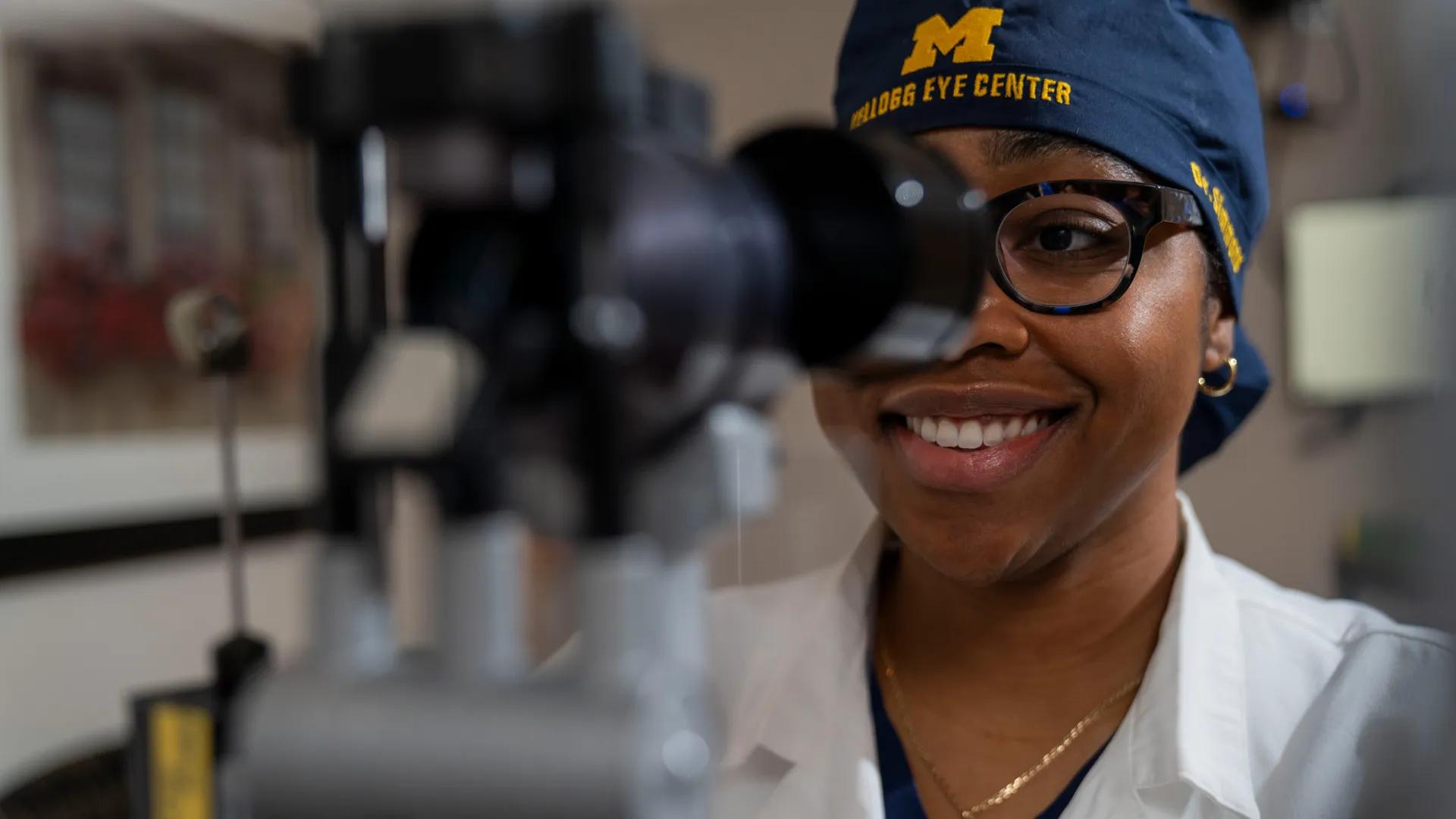 Young black woman wearing glasses and blue Kellogg Eye Center cap looking into optical machine