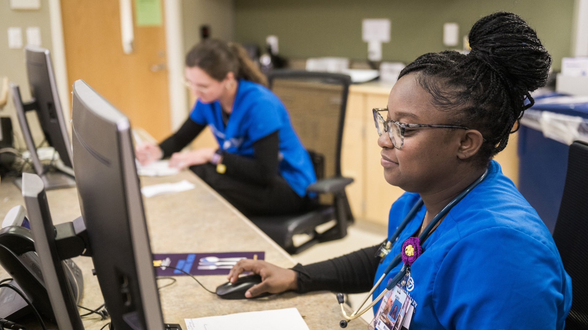 Two U-M Health-West Sparrow nurses seated in front of computers at a workstation. Cupboards and office supplies are visible behind them. 